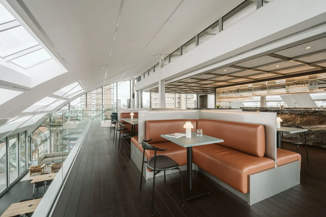 Interior of a modern, stylish restaurant or cafe with booth seating, small tables, and large windows with a city view.