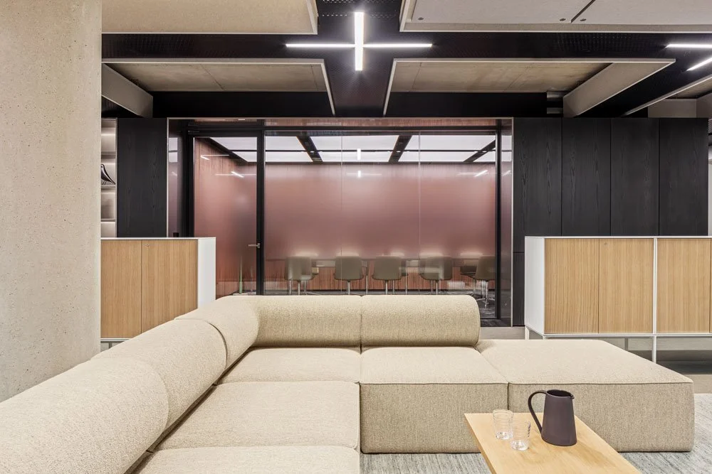 Modern hotel lobby with beige sectional sofa, a wooden coffee table with a black pitcher and glasses, frosted glass wall with bar stools behind, black and wood accent walls, and contemporary lighting fixtures.
