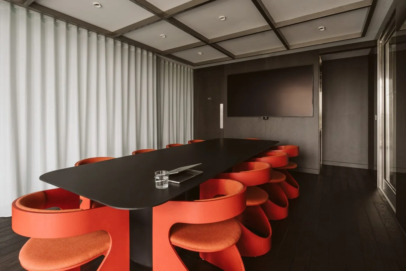A modern conference room with a long black table, red circular chairs, a glass of water, a closed laptop, and a large flat screen TV on the wall. White curtains cover the windows, and the ceiling has recessed lighting.