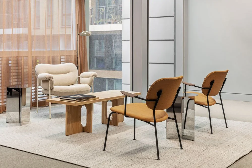 Modern office lounge area with a beige armchair, wooden coffee table, and two orange chairs with black frames.