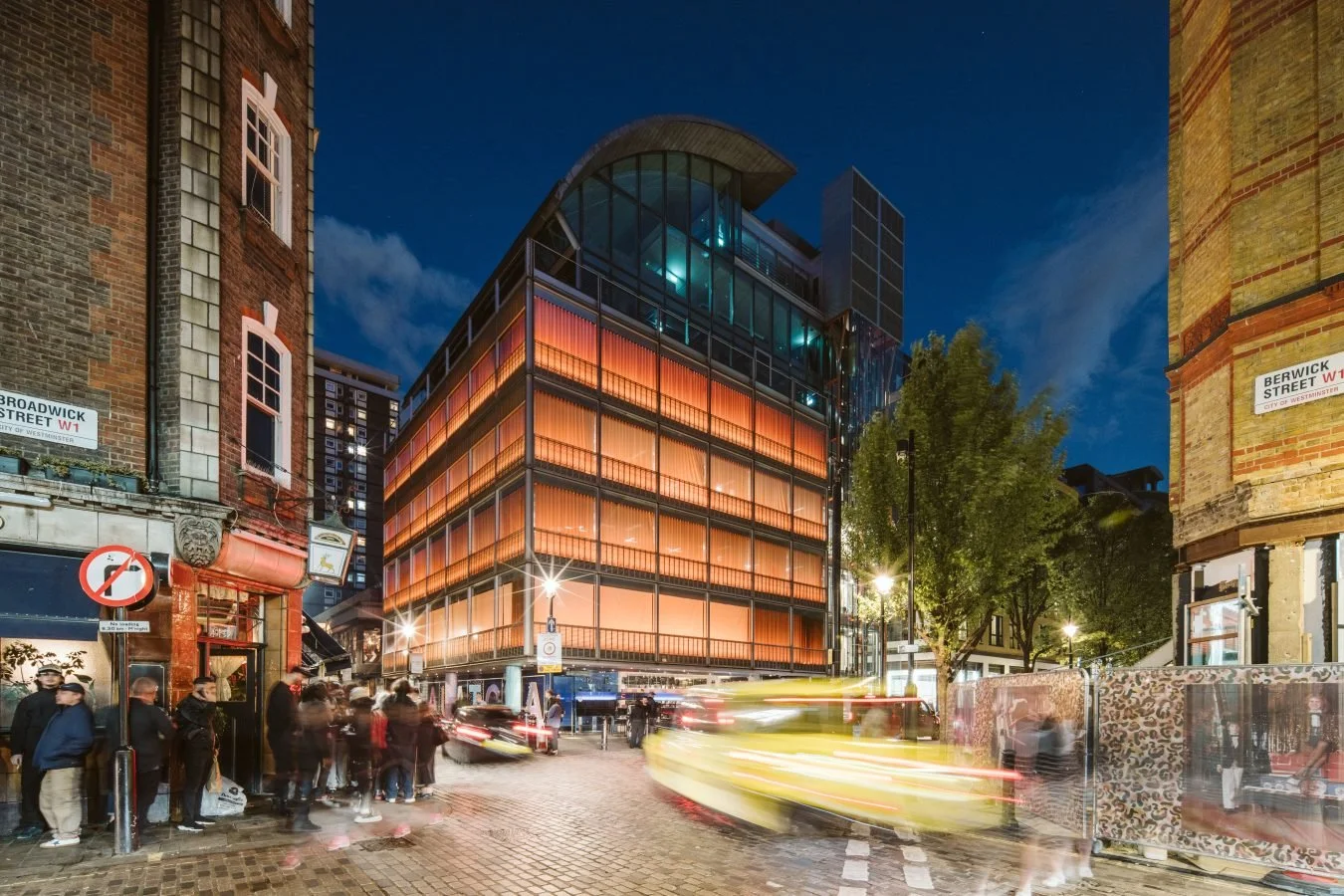 Nighttime city street scene with modern glass building featuring orange lighting, people waiting at a bus stop, and streaks of light from moving vehicles.