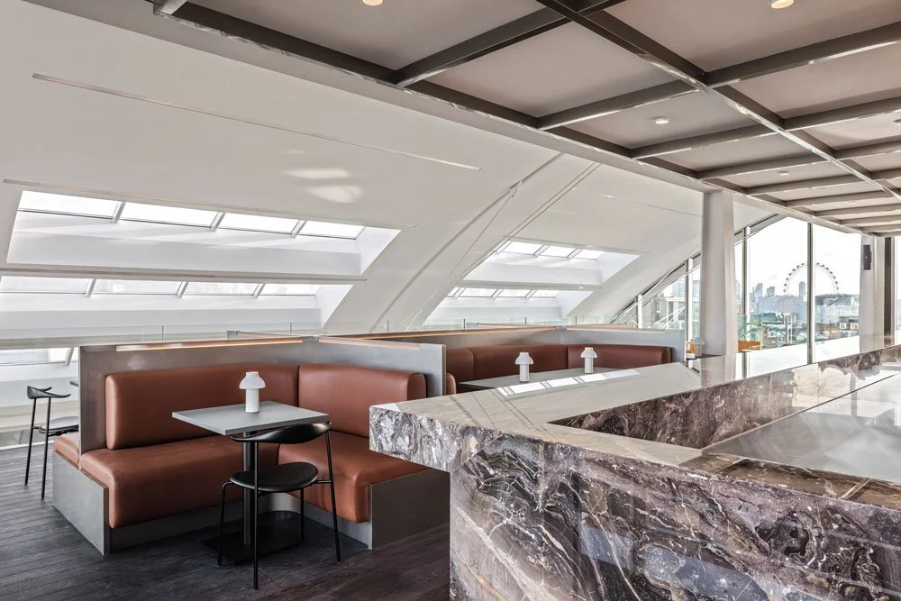 Modern lounge with brown leather booths, small tables with lamps, and large windows showing a cityscape with a Ferris wheel.