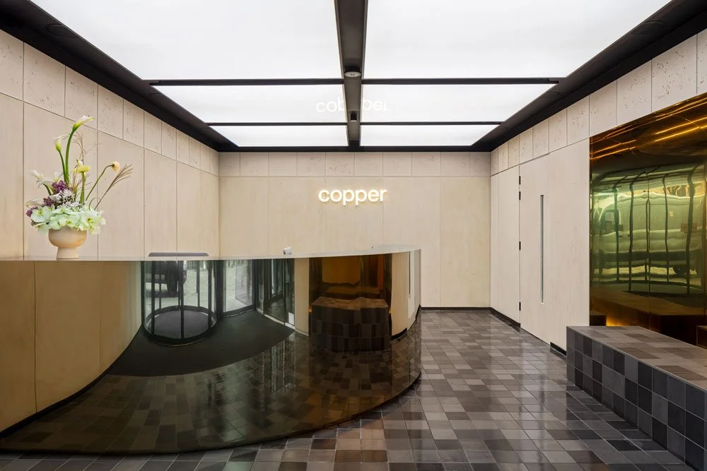 Modern lobby reception area with beige walls, black tiled floor, a curved desk with a gold finish, a white flower arrangement on the desk, and a sign that reads 'copper' on the wall behind the desk.
