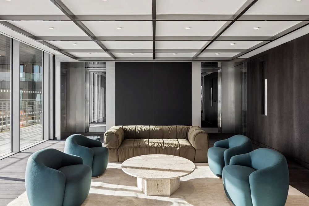 Modern living room with beige sofa, four teal armchairs, a round marble coffee table, large glass sliding doors, and a dark accent wall.