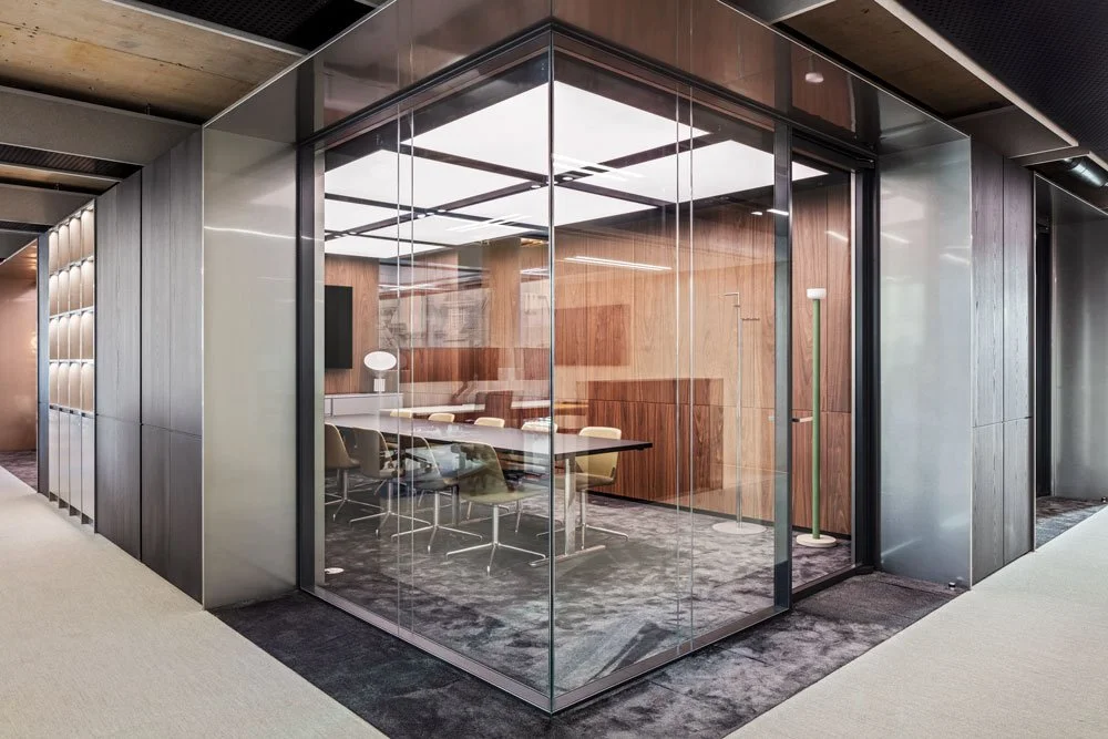 A modern glass-walled conference room with a white rectangular table, beige chairs, and a wooden wall interior, situated in a corporate office space.