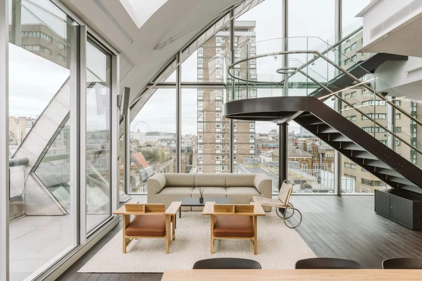 Modern urban living space with large windows, a spiral staircase, beige sofa, and wooden chairs, overlooking city buildings.