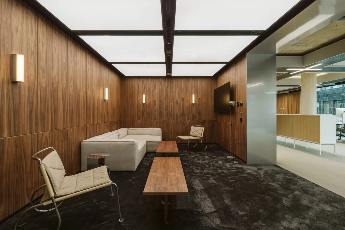 Modern lounge with wooden wall panels, a white sectional sofa, and minimalist chairs, illuminated by wall sconces and ceiling lights.