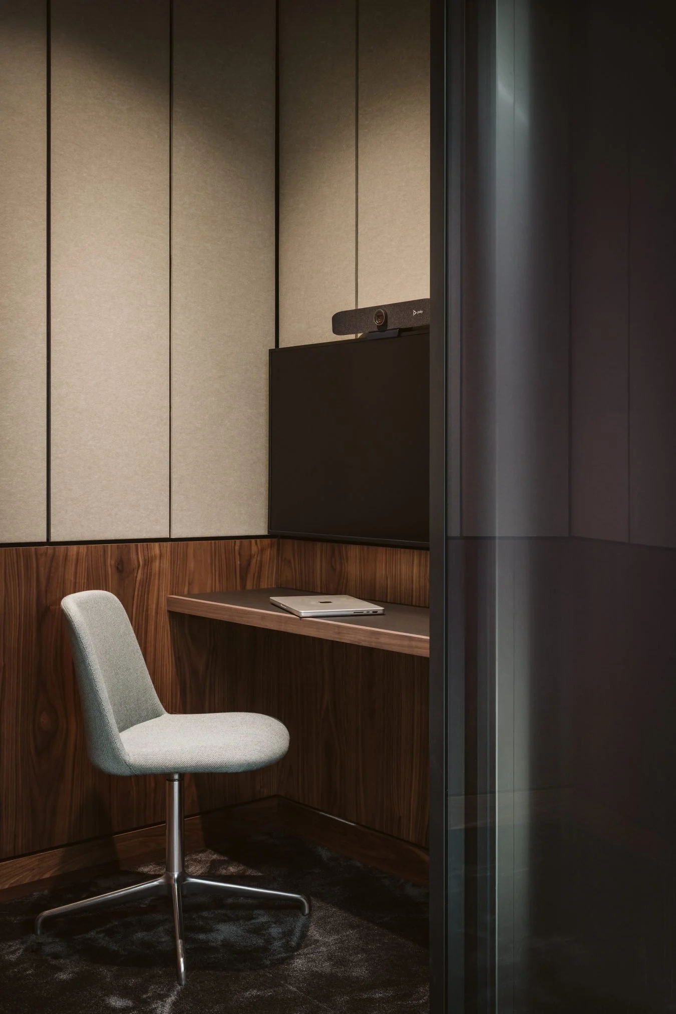 A modern, soundproof Zoom meeting room with beige padded walls, a wood desk, a closed laptop, and a gray upholstered office chair, with a large black monitor and a video conferencing camera on top.