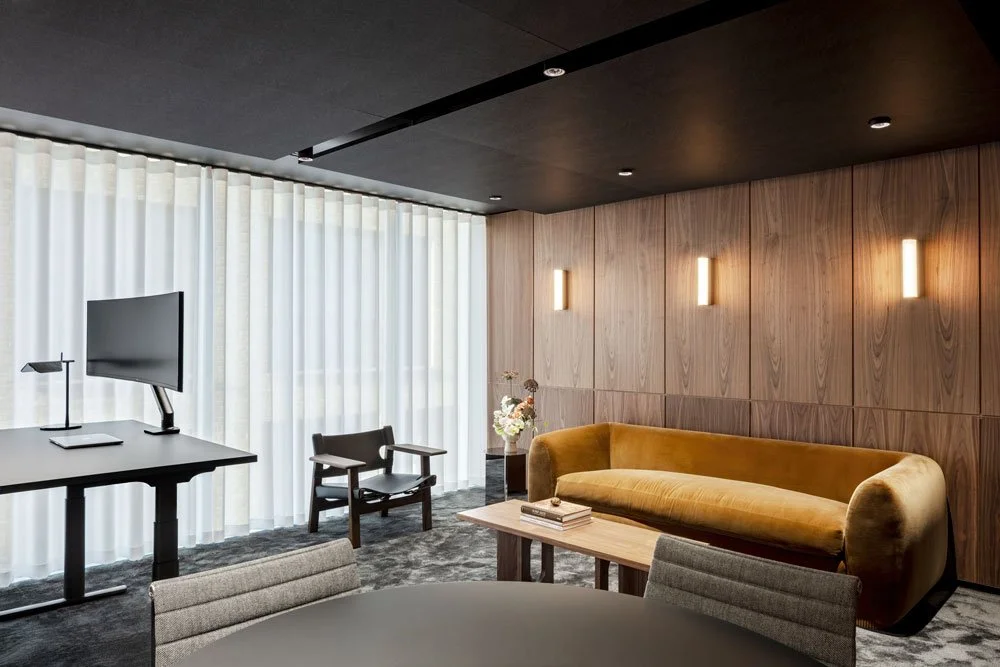 Modern living room with yellow velvet sofa, wooden wall panels, black ceiling, and sheer white curtains, featuring a flat-screen TV, desk with monitor, and a chair.