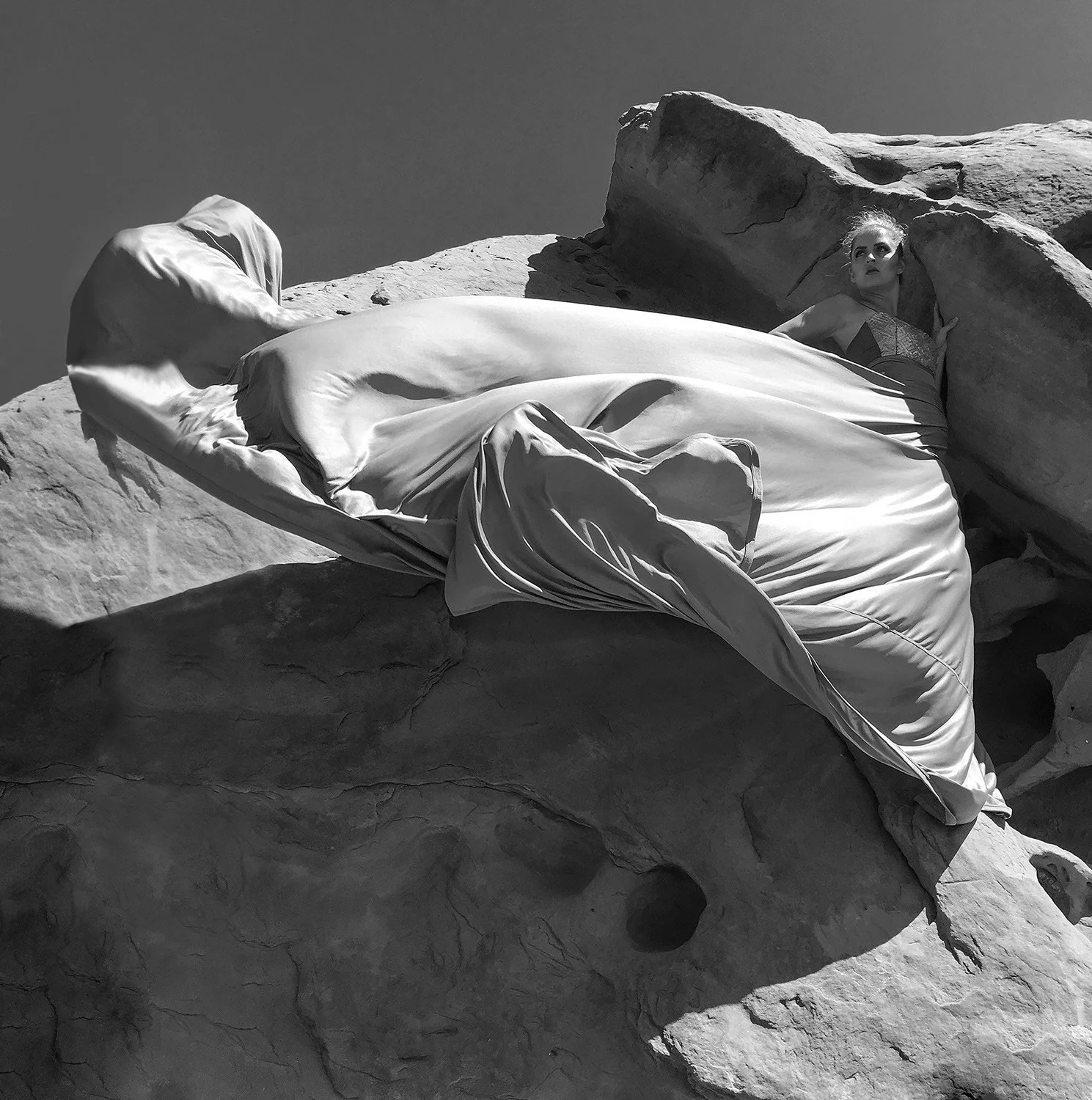 A woman in a satin gown lying on a large rock formation outdoors, with her head resting against a smaller rock, in a black and white photograph.