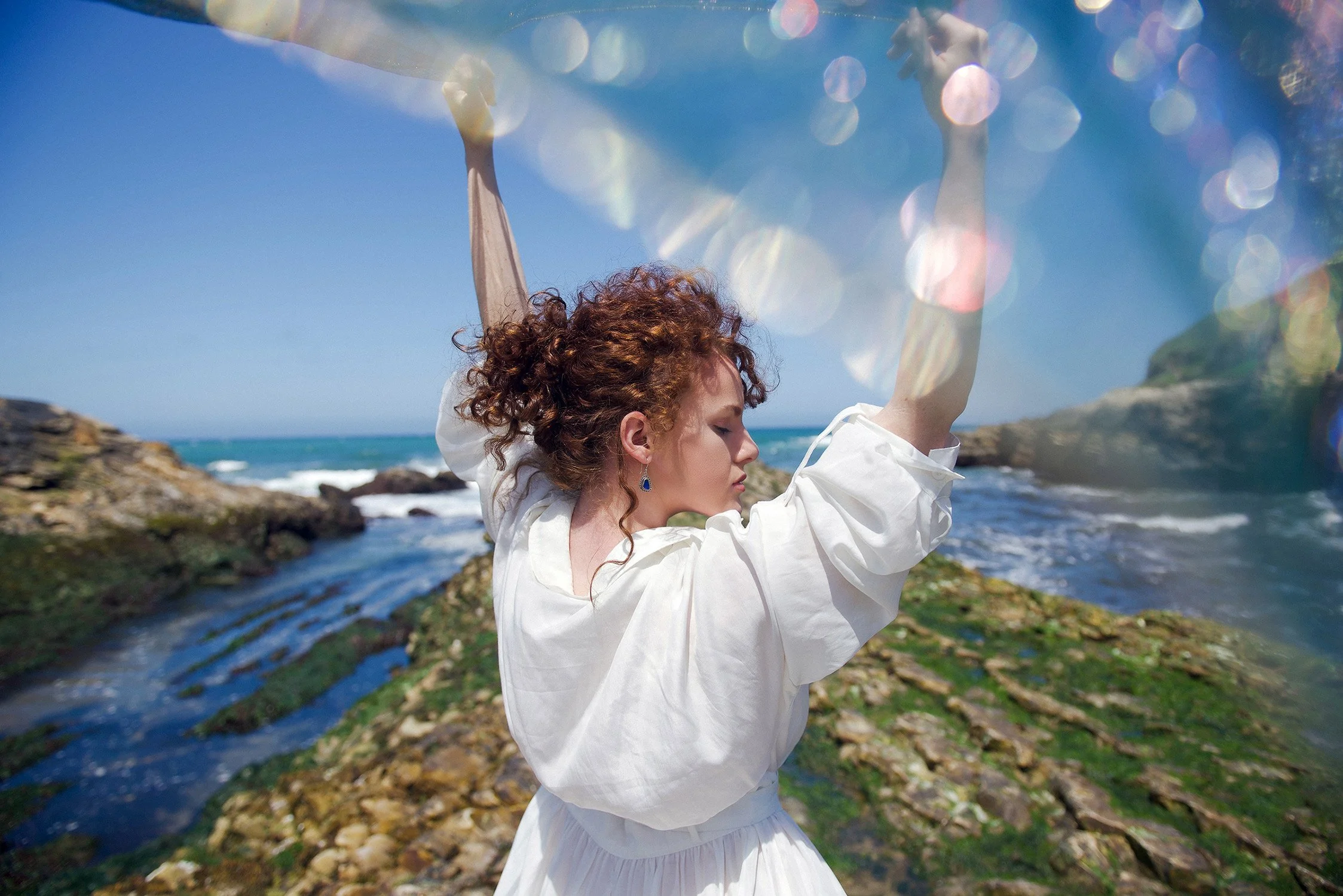 A woman stands at the waters edge holding a shimmering scarf
