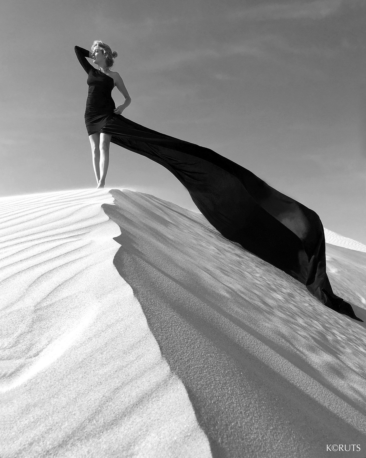 A woman stands poised on a white dune in an elongated black dress