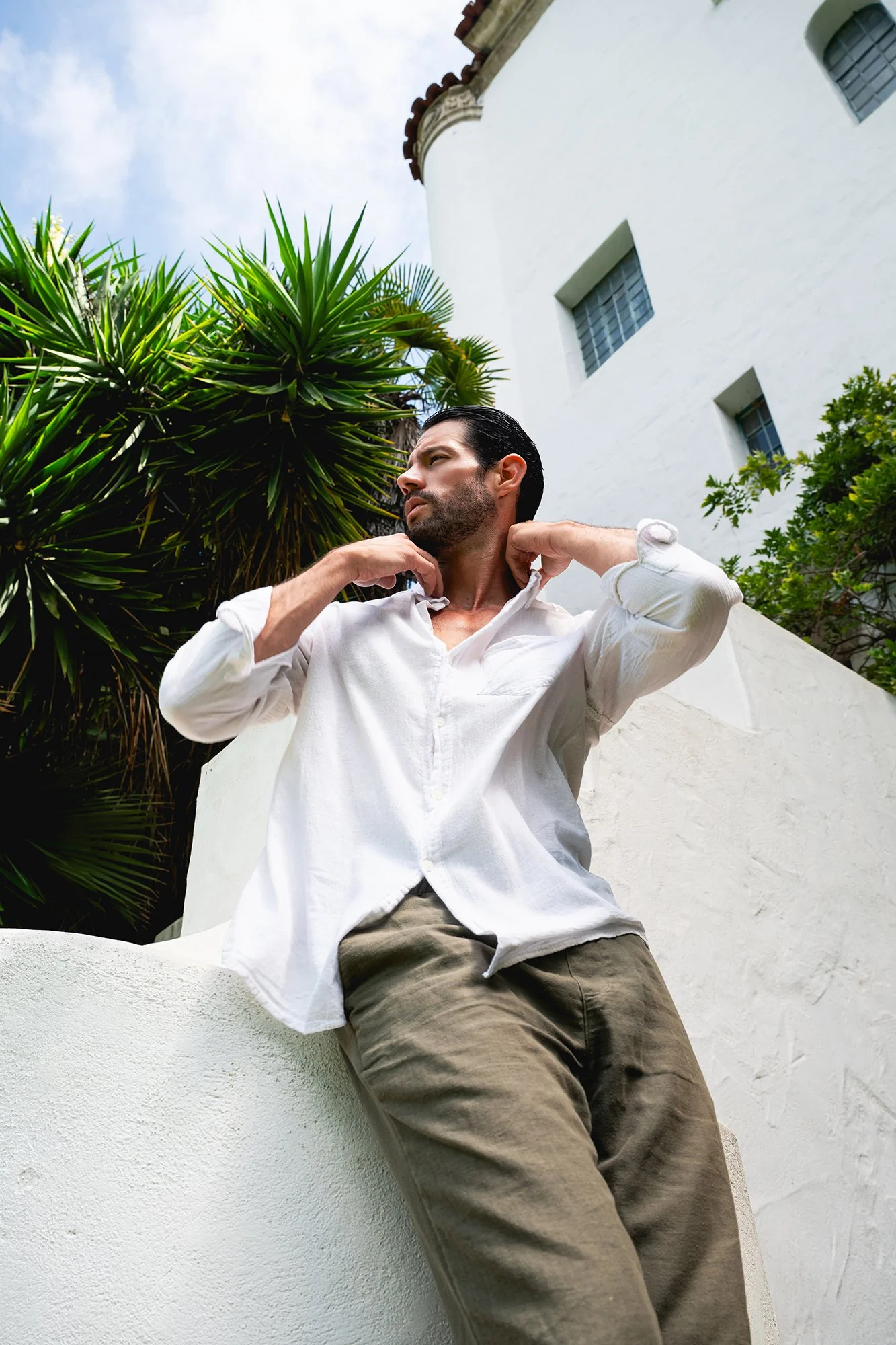 Editorial menswear photography featuring a Spanish male model in casual linen apparel leaning against a courtyard archway in traditional Spanish architecture.
