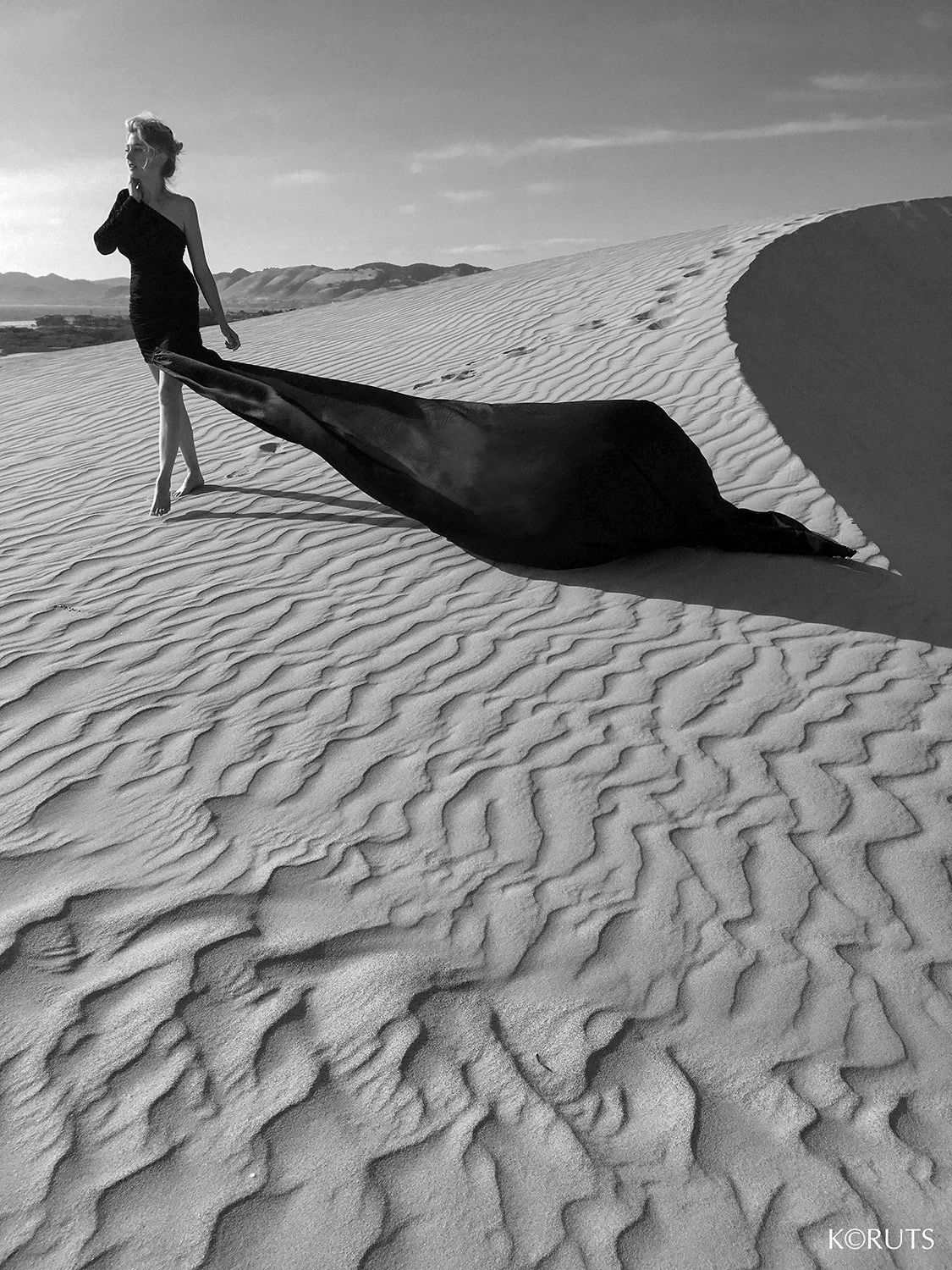 a woman in a black dress walks out of the sand dunes