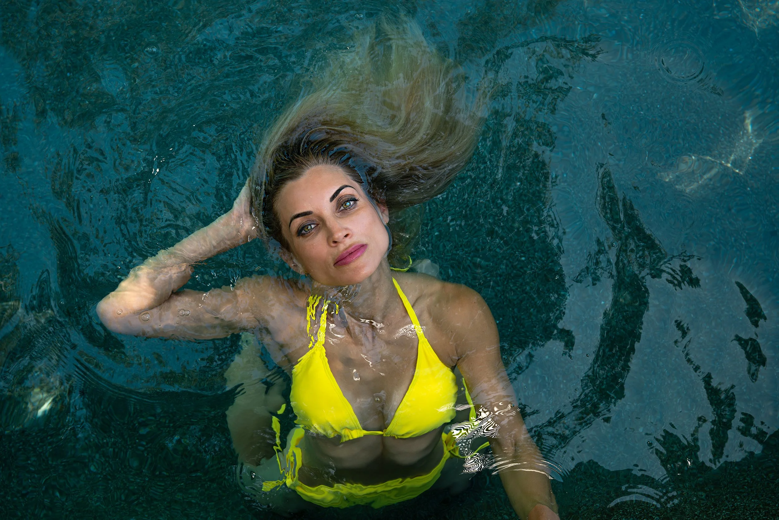 An overhead aerial portrait of a woman wearing a bright yellow swimsuit floating in a vibrant blue swimming pool, styled for a commercial lifestyle photography portfolio.