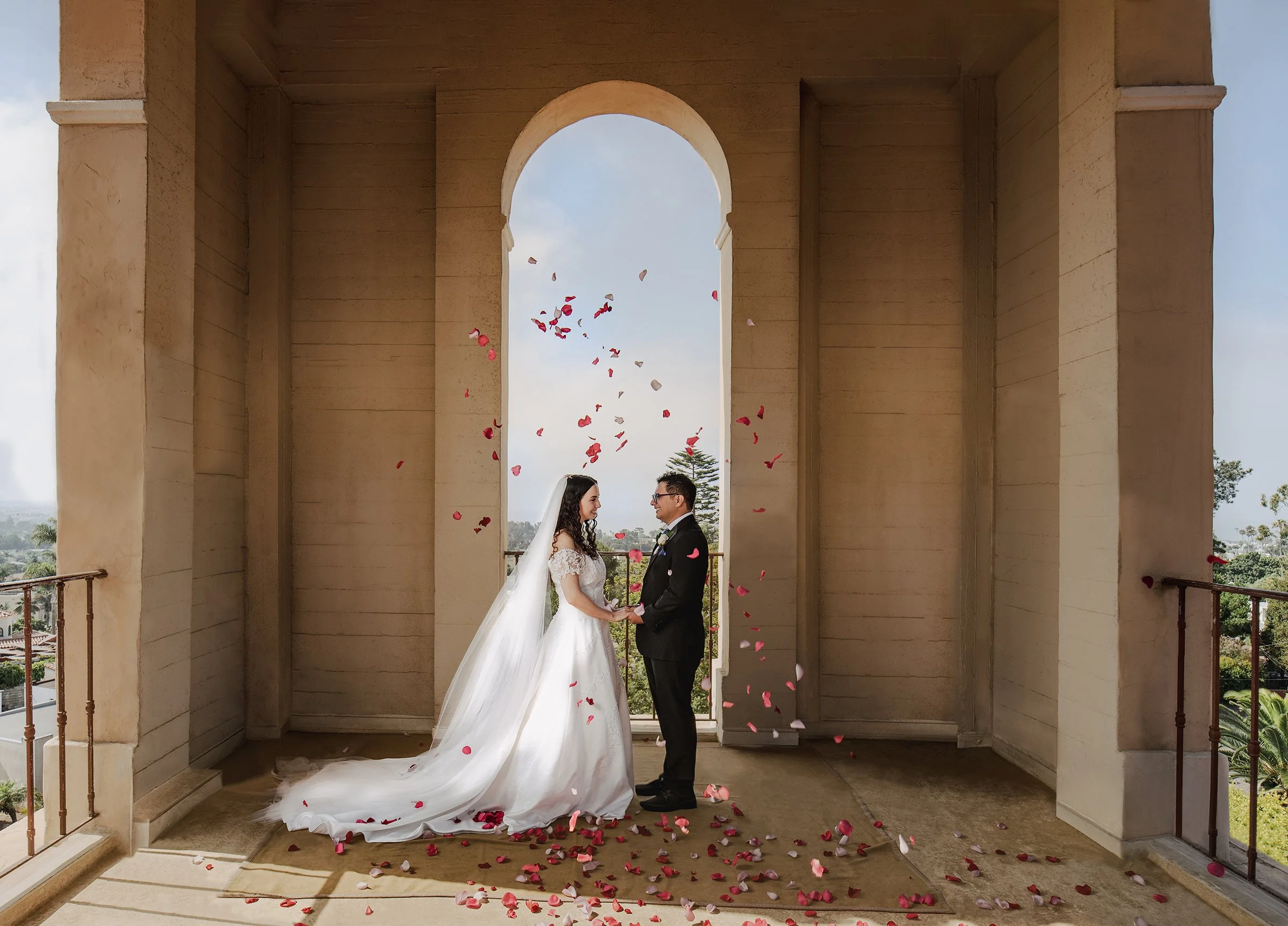 A couple stand under falling rose pedals in the bell tower of the local church.