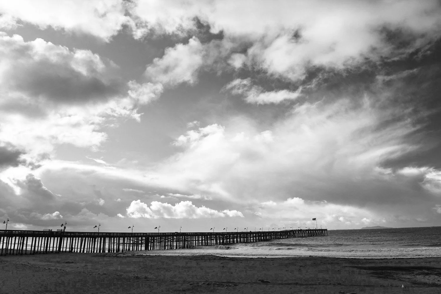 Black and white photo of a Ventura pier from a distance with the sky and cloud spanning the image