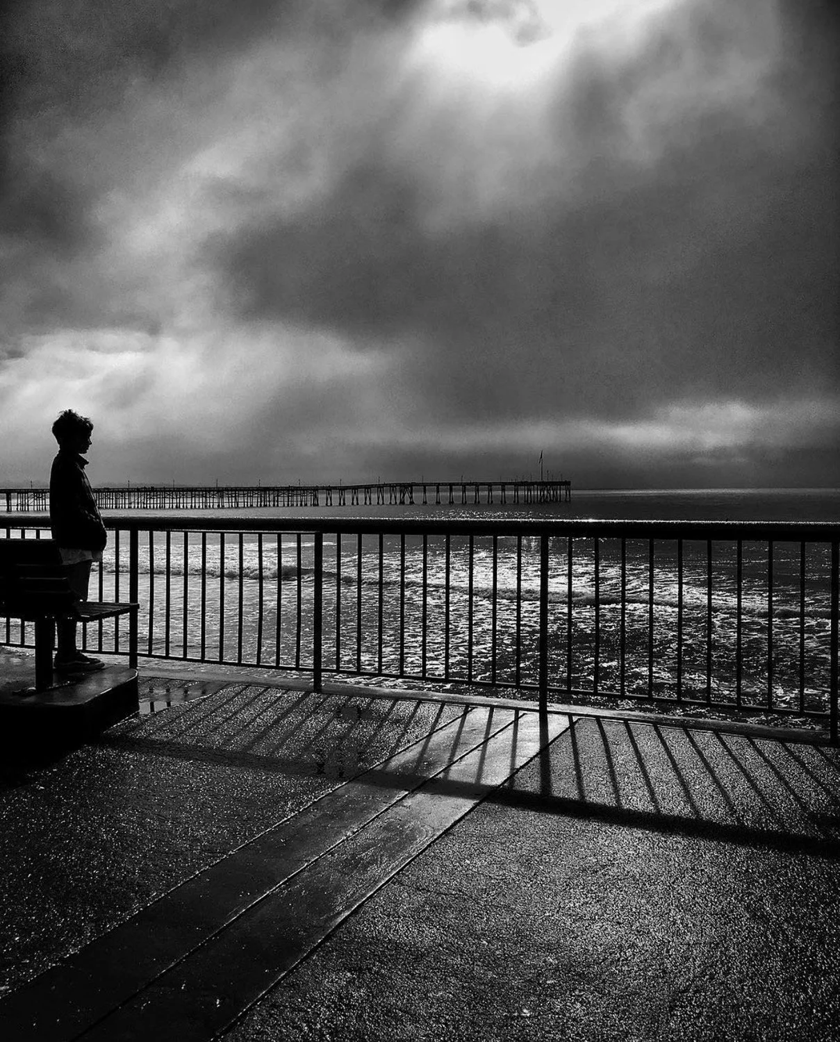 Black and white photo of a child looking out from the pier in Ventura California after a storm has passed, the clouds have parted and the sun is shining through