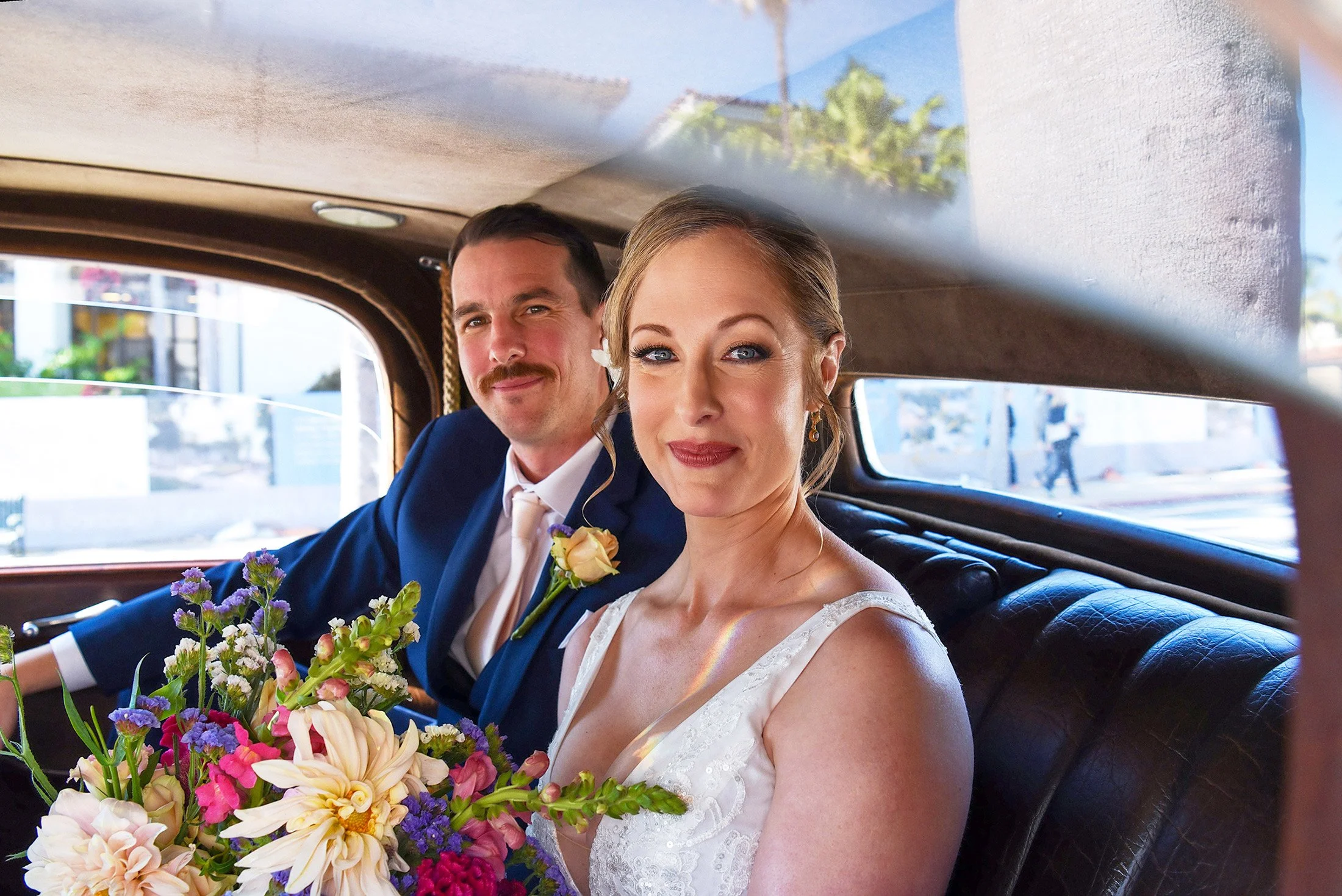A happy couple smiles from the back seat with flowers in the brides hands