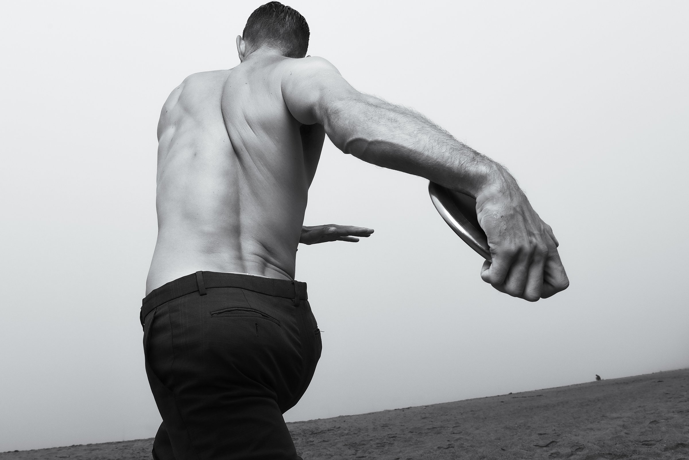A black-and-white photo of a shirtless man holding a frisbee on a beach, viewed from behind.