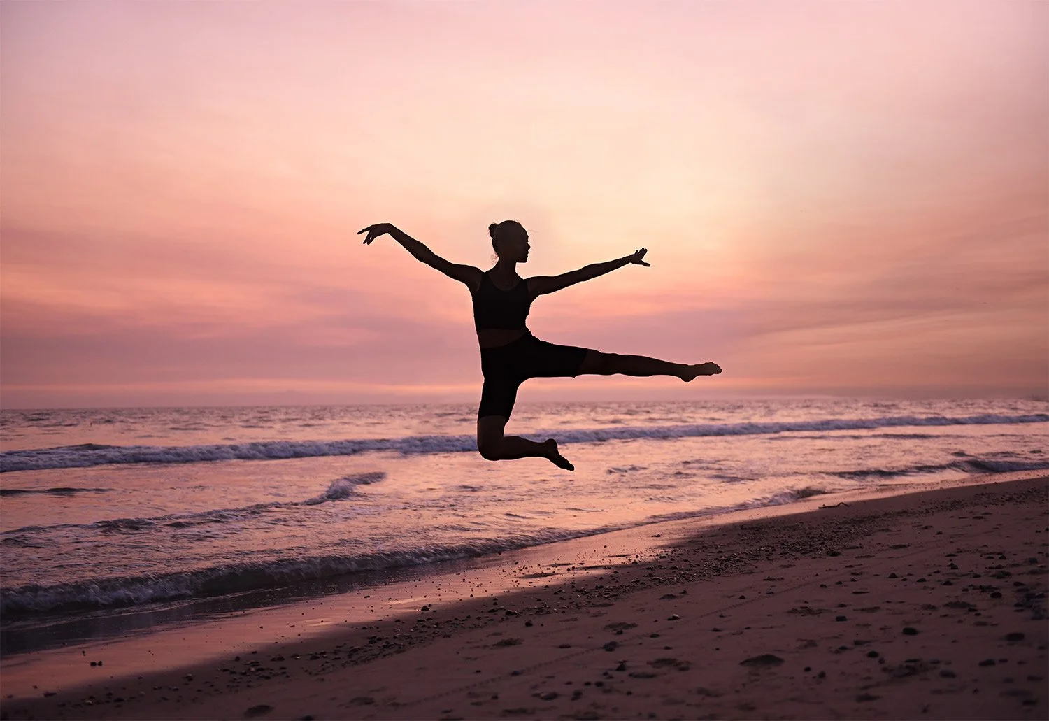 Woman ballet dances and jumps on the beach with a purple sunset in Ventura California.