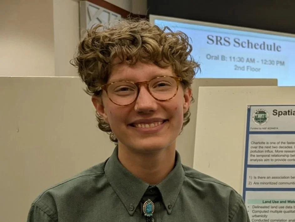 Closeup of Dezzie wears glasses and a gray dress shirt, they are smiling at a science research poster presentation for the 2025 summer OUR conference.