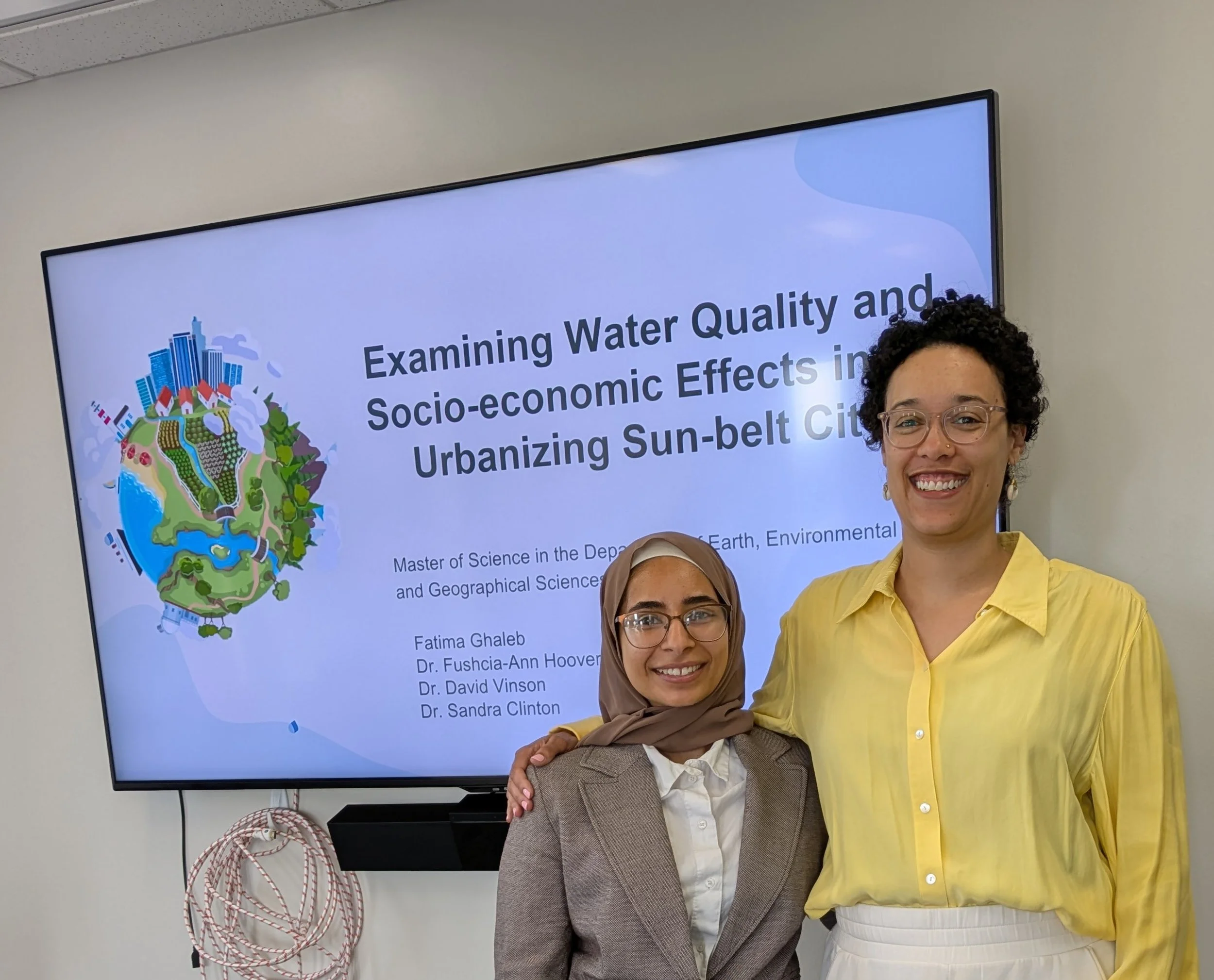 Dr. Hoover wears glasses and a yellow shirt, and Fatima wears a hijab and a beige blazer with a white blouse. They both smile and pose in front of a presentation for Fatima's thesis defense.