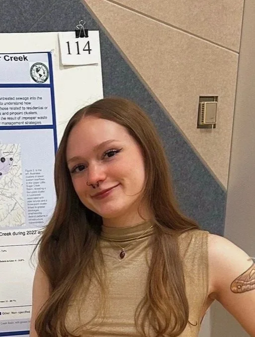 Grace wearing a beige top and with red hair, smiles in front of a poster for the spring 2025 OUR conference.