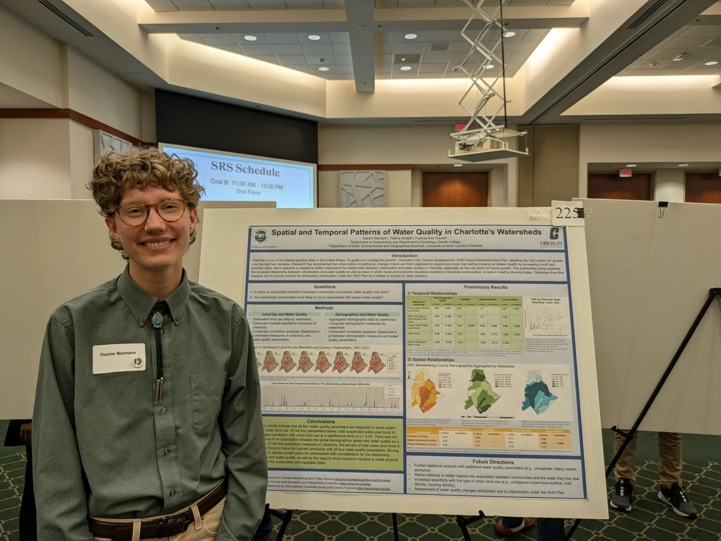 Dezzie wears glasses and a gray dress shirt, they are smiling at a science research poster presentation for the 2025 summer OUR conference.