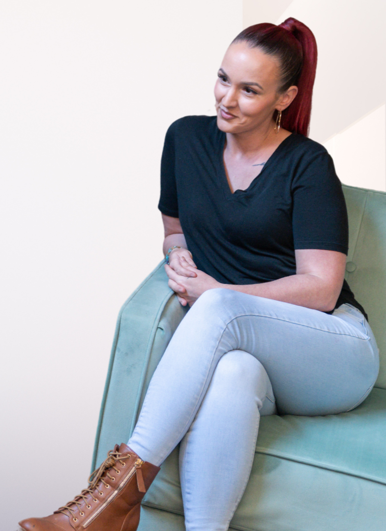 A woman with long red hair, wearing a black t-shirt, light blue jeans, and brown ankle boots, sitting on a light green sofa with her hands clasped on her lap, smiling softly.