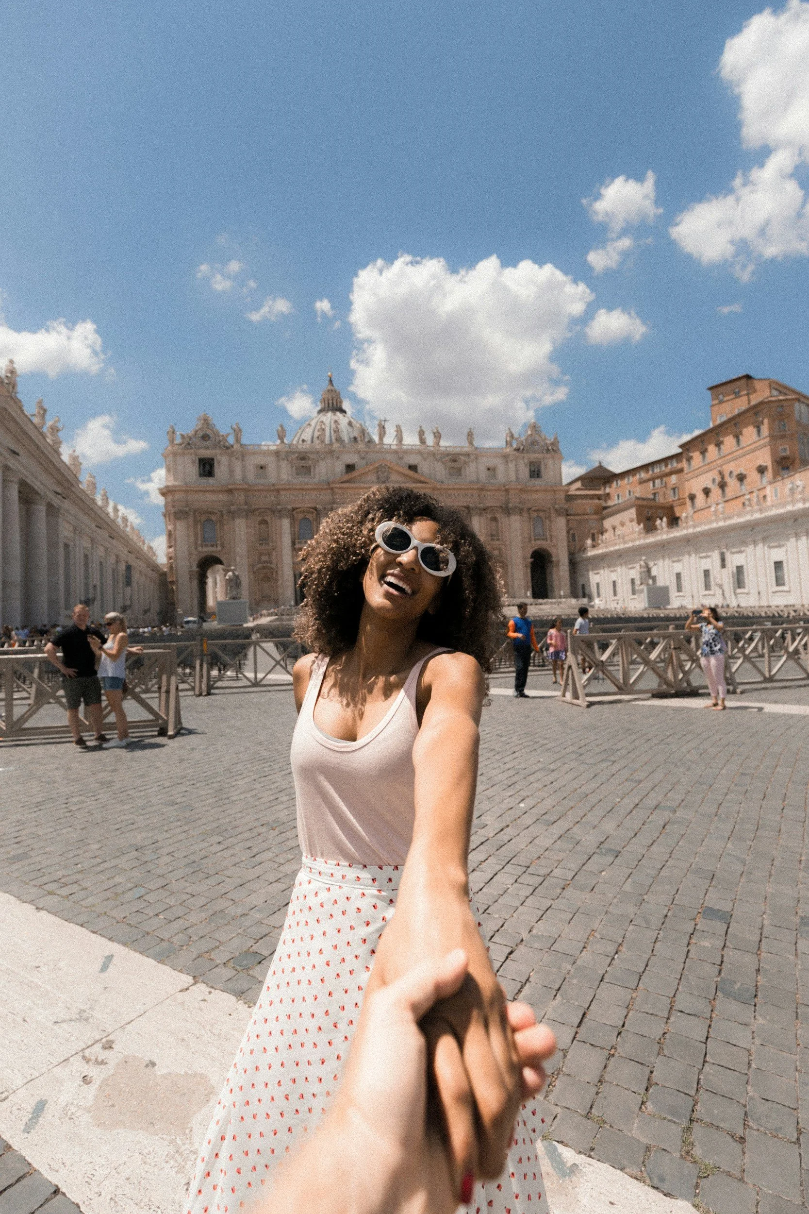 A woman with curly hair, wearing sunglasses, a beige top, and a white skirt with red polka dots, smiling and holding hands with the person taking the photo in front of St. Peter's Basilica in Vatican City on a sunny day.