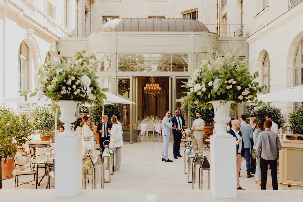 Un groupe de personnes bien habillées se tient dans une cour élégante avec des plantes en pot et de grandes compositions florales. L'arrière-plan montre une structure en verre avec un chandelier, indiquant une réception ou un événement social en exté
