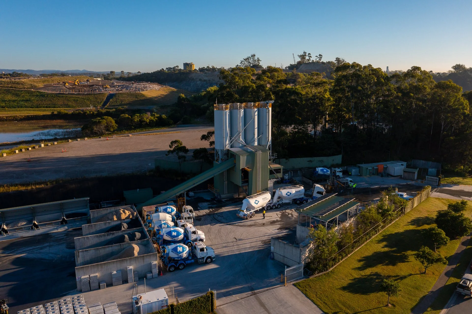 Industrial facility with cement silos, concrete mixers, trucks, and storage containers, set against a landscape with trees, a body of water, and hills in the background.