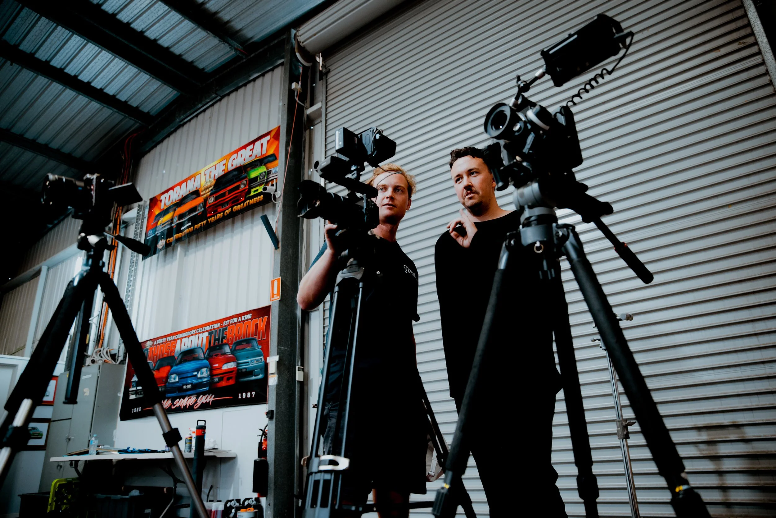 Two men talk and examine camera equipment in a garage with car posters on the wall.