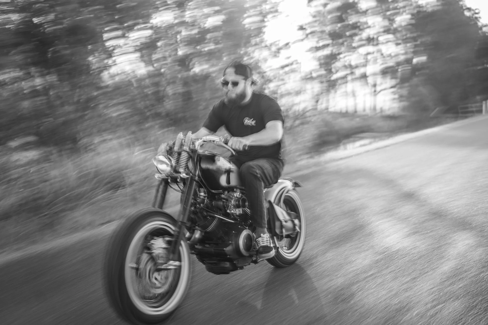 A man riding a motorcycle on a road surrounded by trees, captured in motion with a blurred background in black and white.