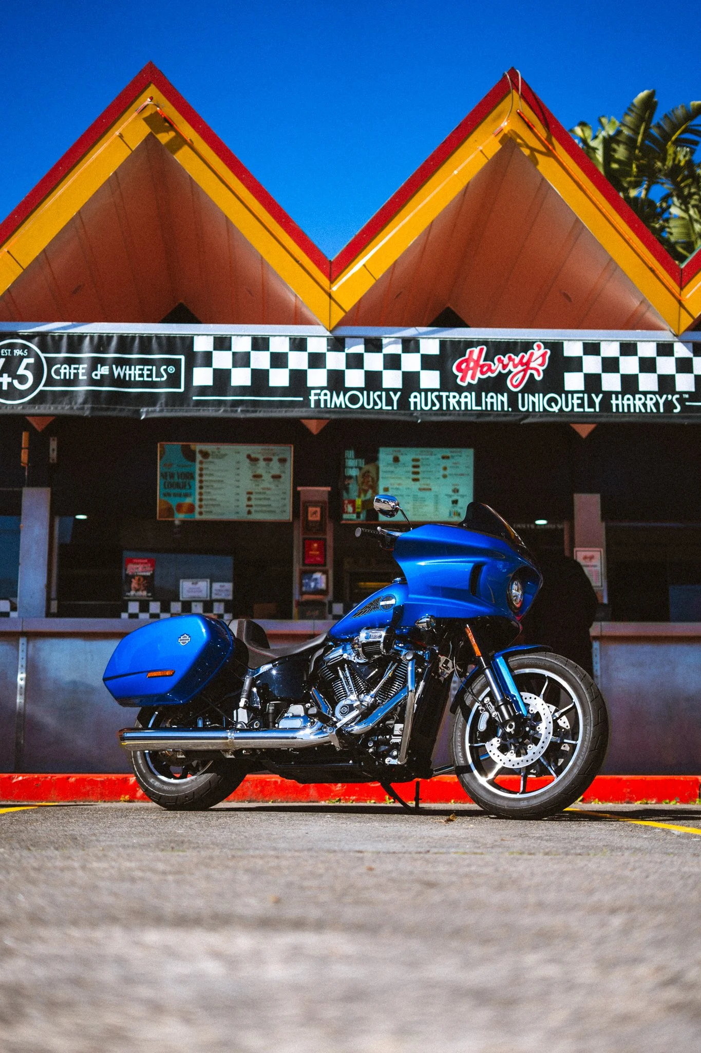 A blue motorcycle parked in front of a food stand at Harry's Café de Wheels, with a checkered flag design on the sign and a colorful roof background. Harley Davidson Australia Low Rider ST