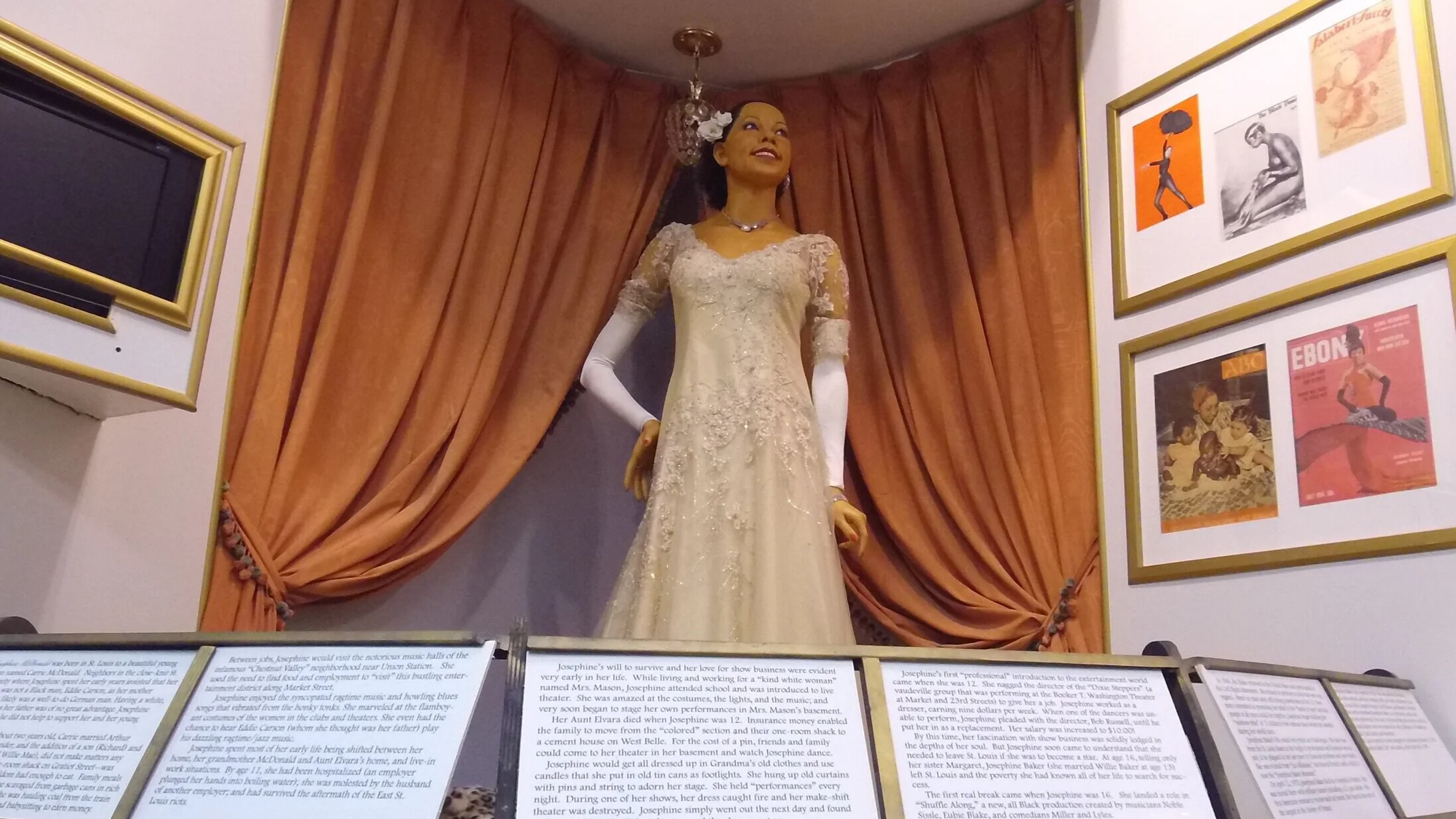 A mannequin dressed in a vintage cream-colored wedding gown with lace applique, displayed in front of orange curtains, surrounded by framed artworks and informational plaques.