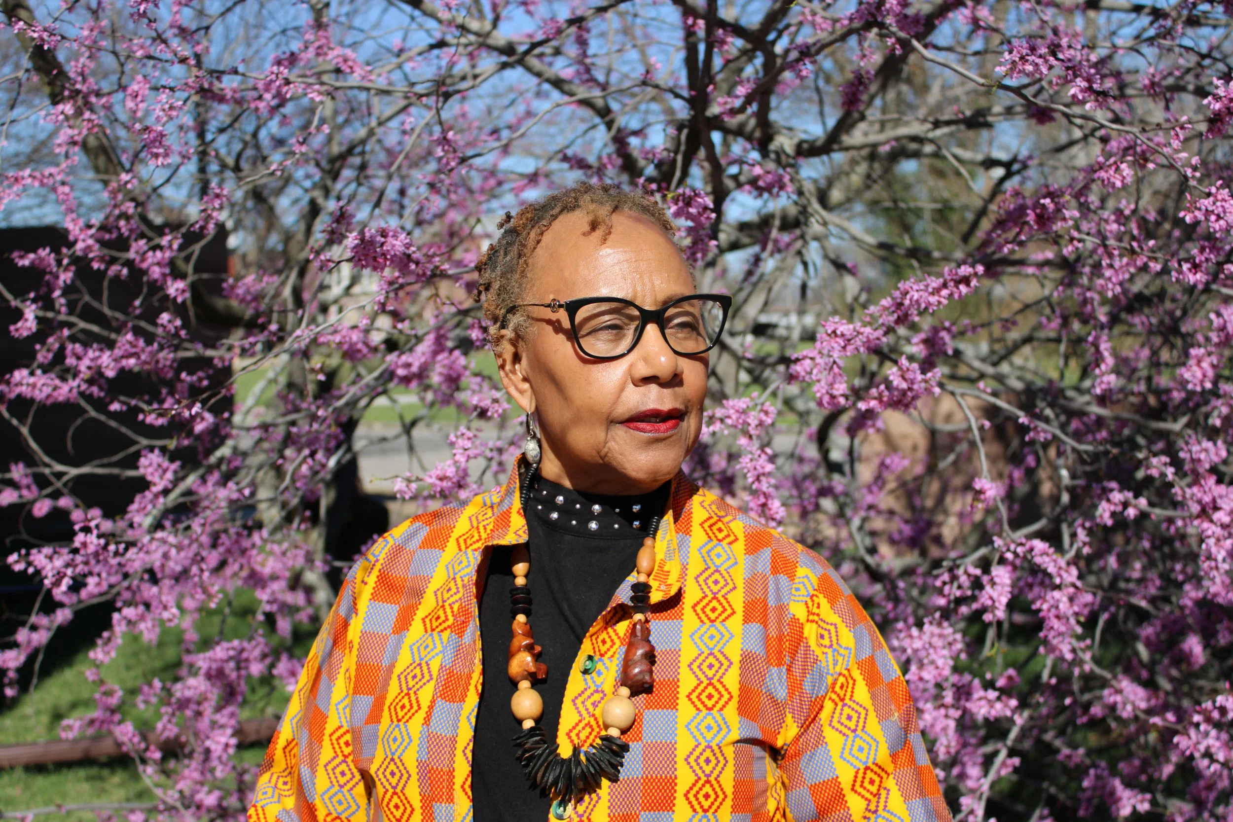 Lois Conley with glasses and earrings stands outdoors in front of purple blooming trees, wearing colorful patterned clothing and a necklace.