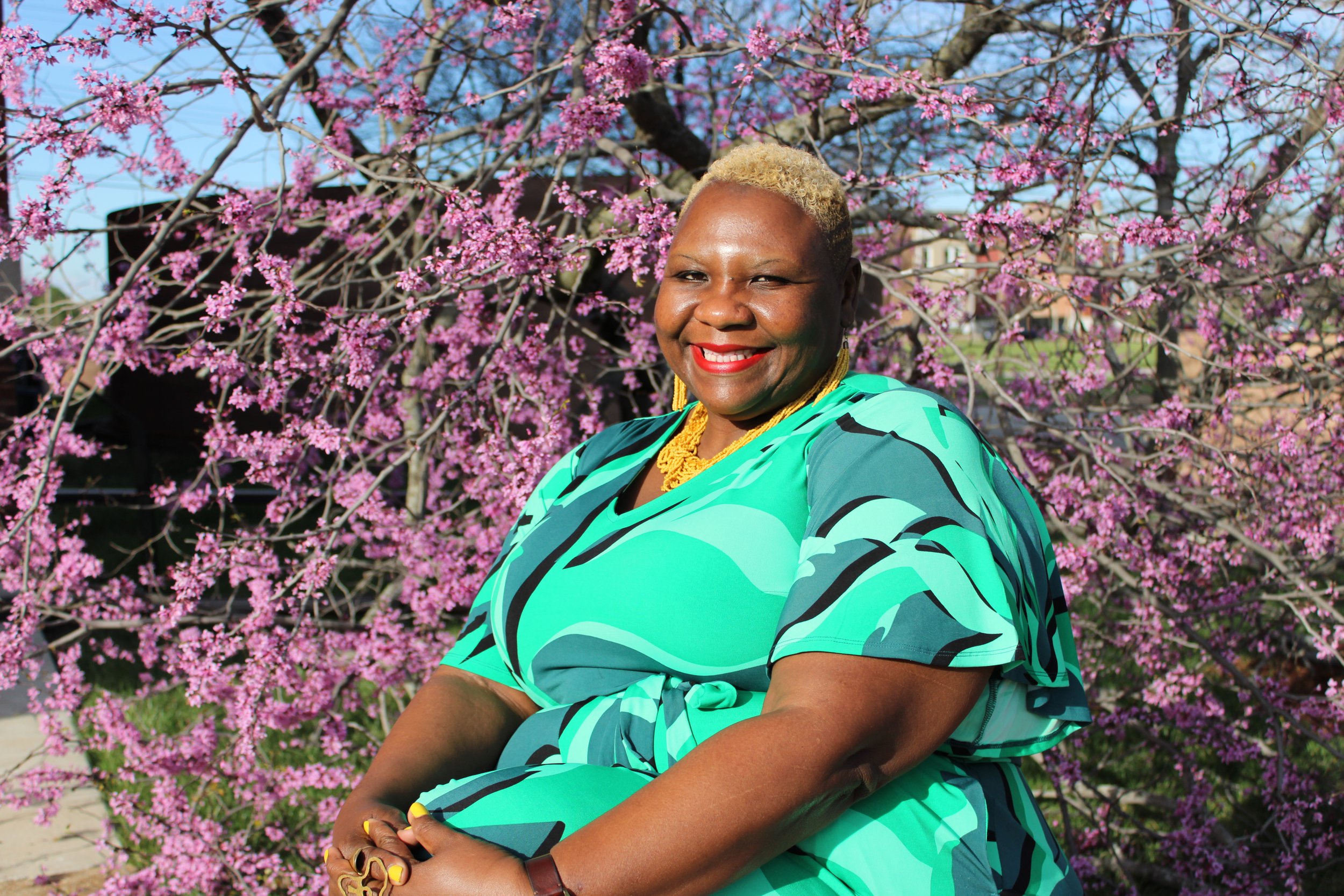 Yolanda Williams sitting outdoors in front of a blooming pink flowering tree, wearing a teal and black patterned dress with gold jewelry, smiling and looking at the camera.