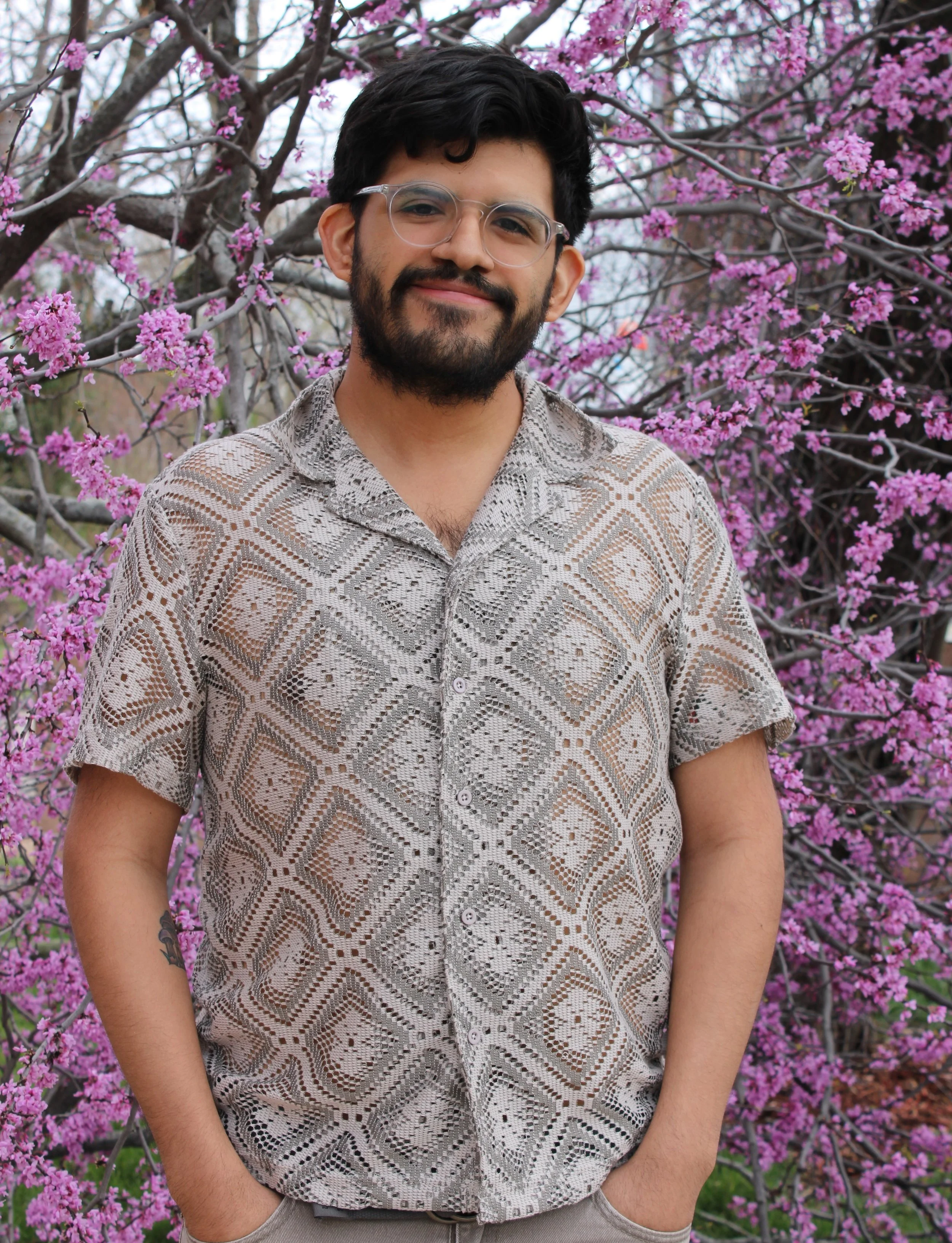 Zeles Vargas with dark hair, beard, and glasses, smiling while standing outdoors in front of a tree with pink blossoms. He is wearing a patterned beige and white button-up shirt with short sleeves.