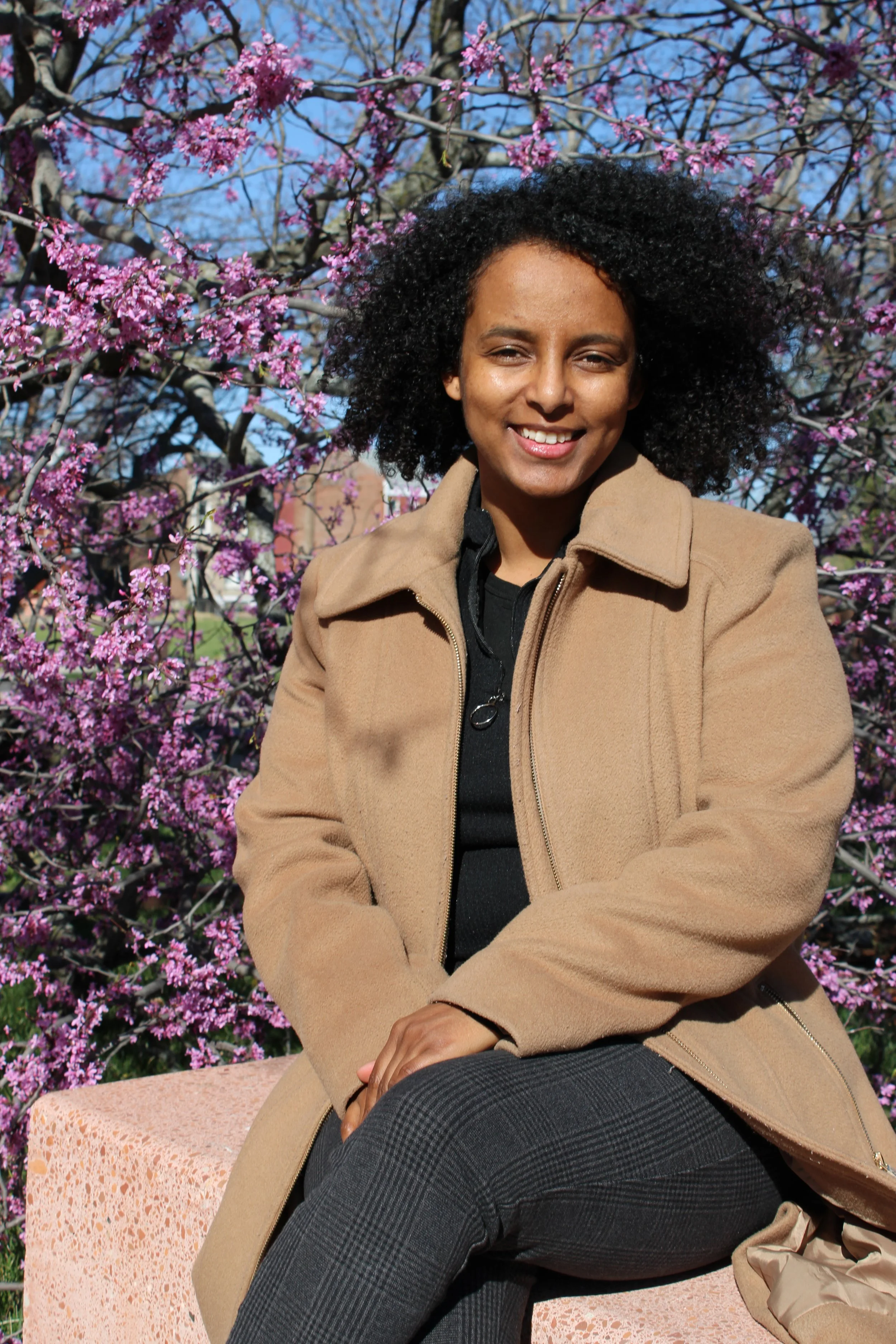 A Sewasew Assefa sitting on a bench outdoors, smiling, with purple flowering trees behind her on a sunny day.