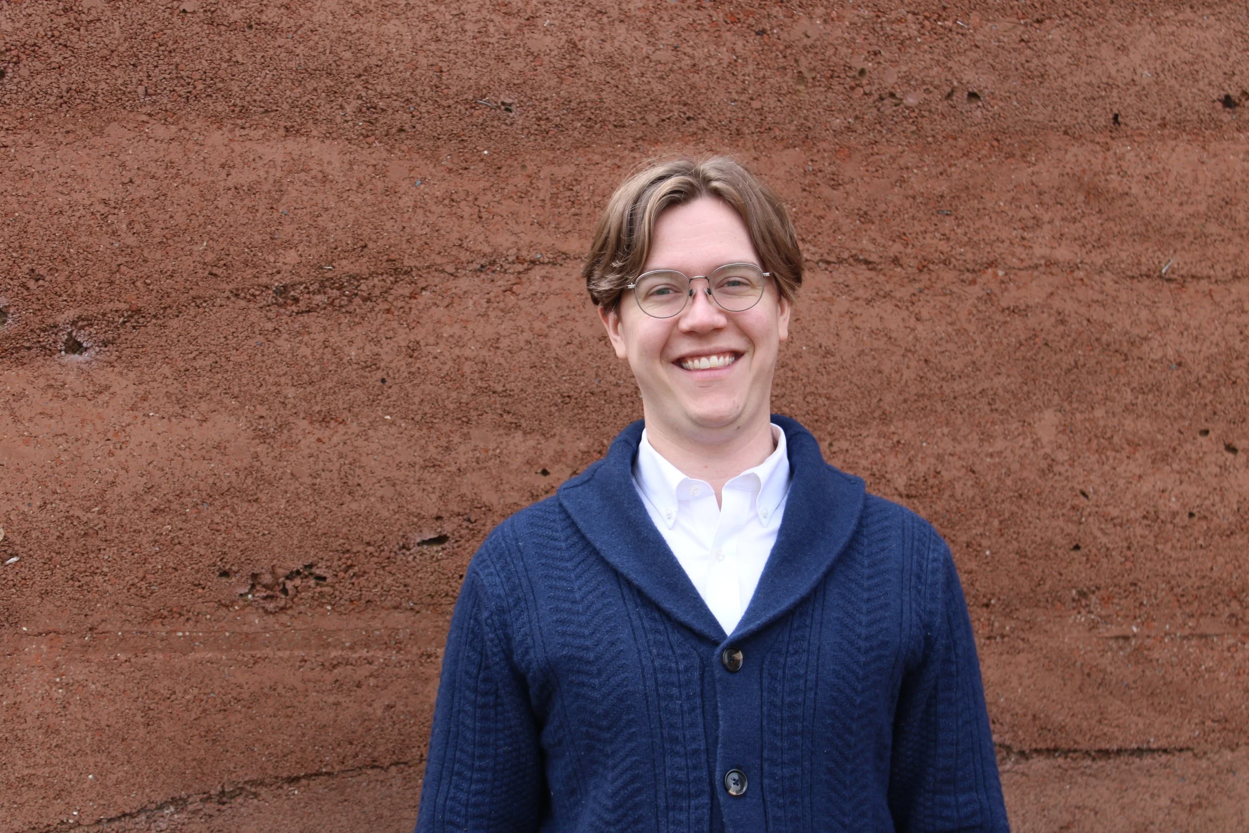 Jonah Lynd-Porter with glasses and brown hair, wearing a white shirt and a blue cardigan, standing against a red brick wall.
