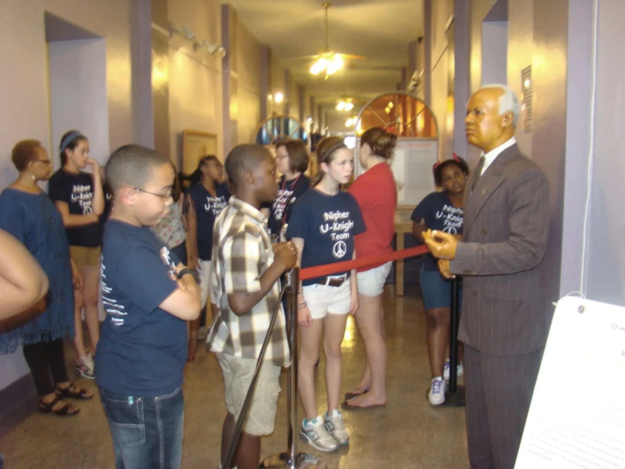 A group of children, mostly wearing matching U-Knight Team shirts, listening to a man in a suit speaking during a museum or exhibit tour, with some adults present in the background.