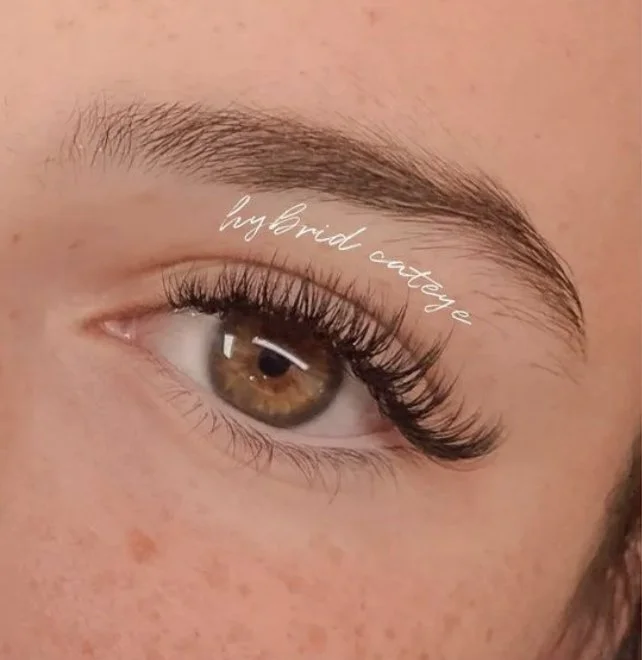 Close-up of a person's eye with makeup, showing brown iris, long eyelash, and eyebrow. Text over the eyelid reads 'hybrid couture'.