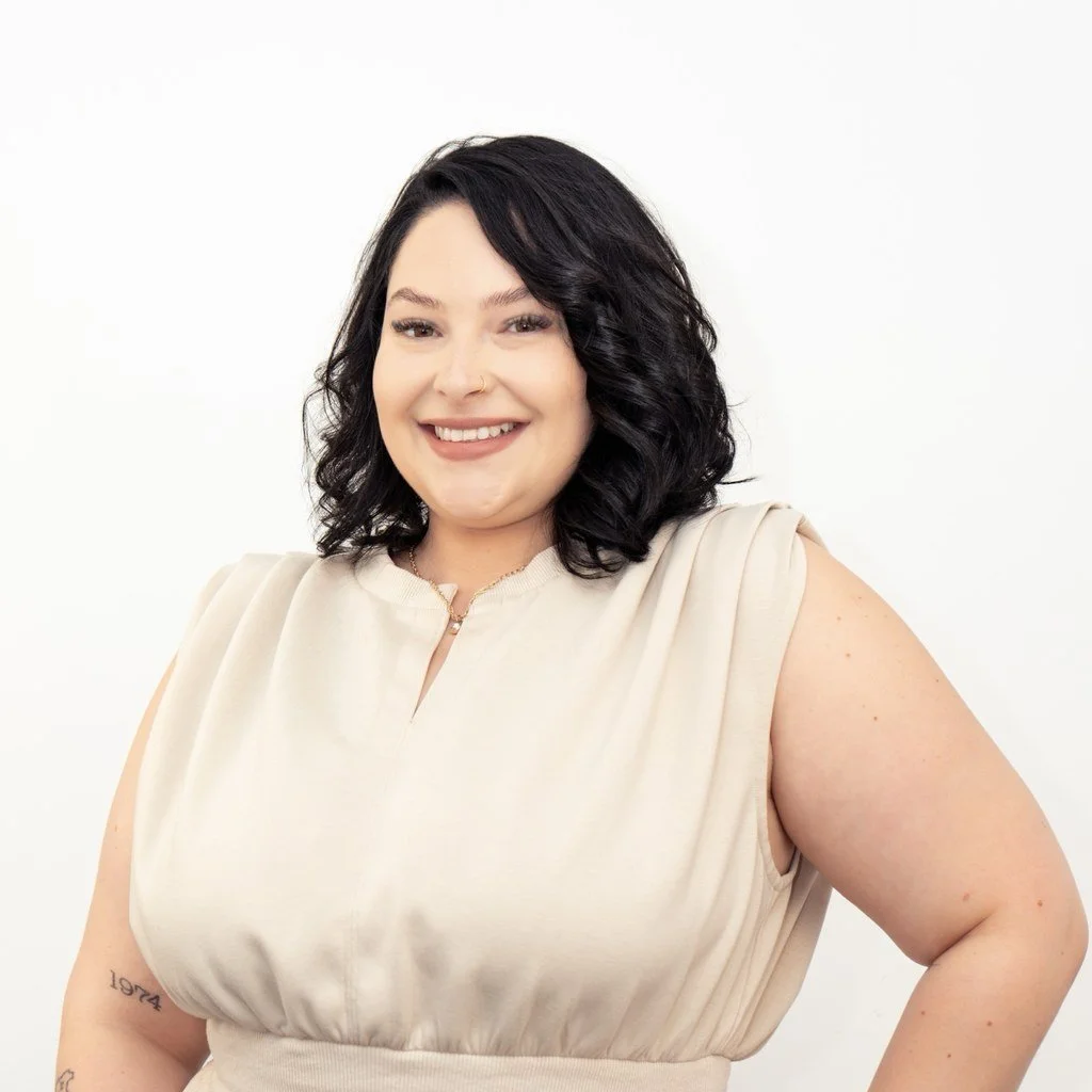A woman with long black hair and light skin, wearing a black lace top and dark jacket, holding a red and black tool, sitting on a stool against a plain gray background, smiling at the camera.