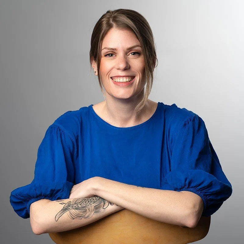 A woman in a blue blouse smiling and leaning on a wooden chair, showcasing a tattoo on her forearm.