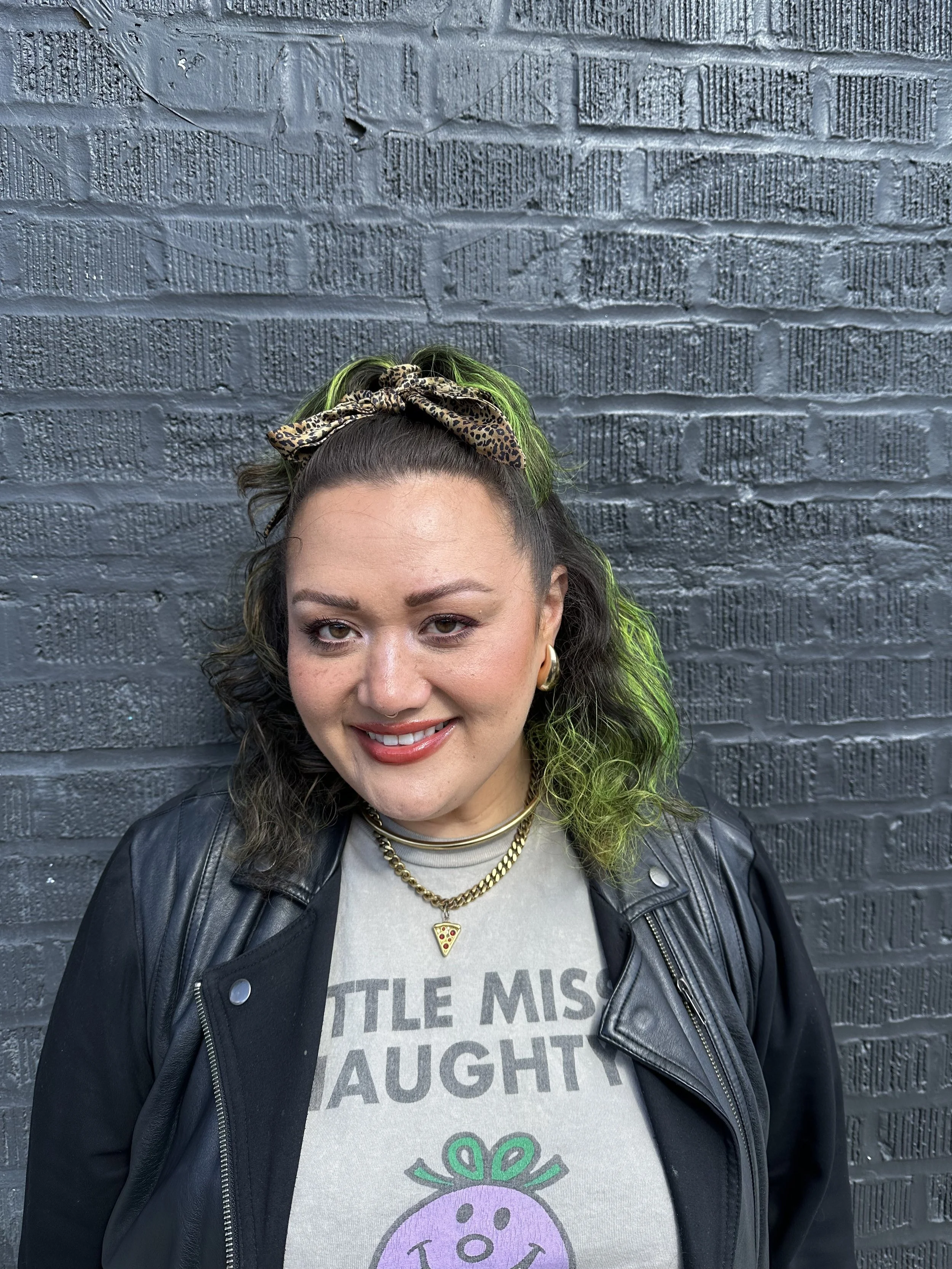 A woman with dark wavy hair highlighted with green, wearing a leopard-print hair scarf, a black leather jacket, and a graphic t-shirt with a smiling cartoon vegetable, standing in front of a textured dark gray brick wall.