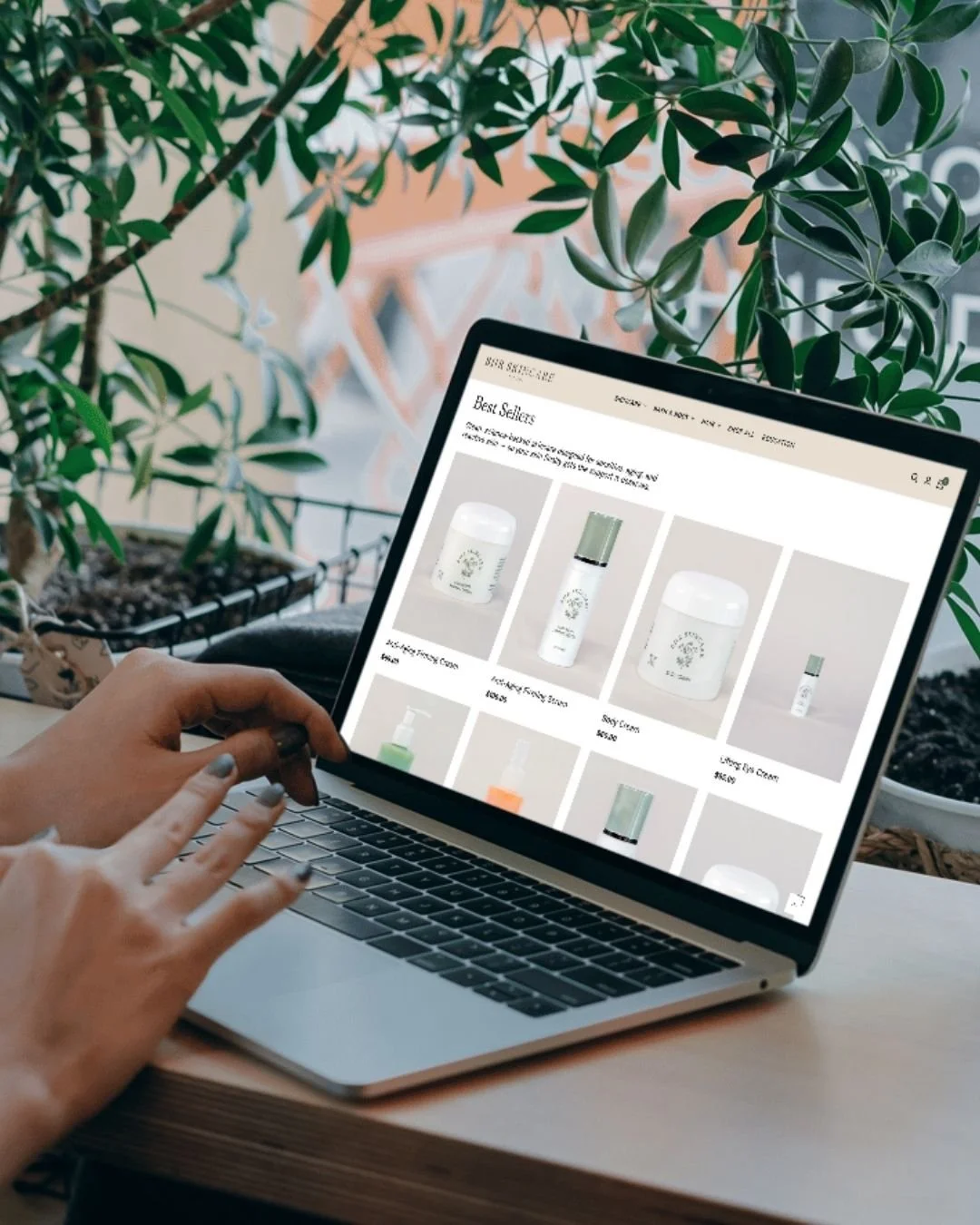 Hands over a laptop on a wooden counter with a lush green plant in the background and a collection of skincare products on the laptop screen