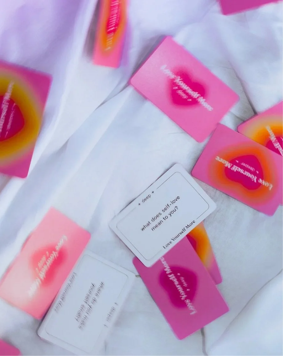 Pink and orange "love yourself more" card deck cards strewn across a white blanket
