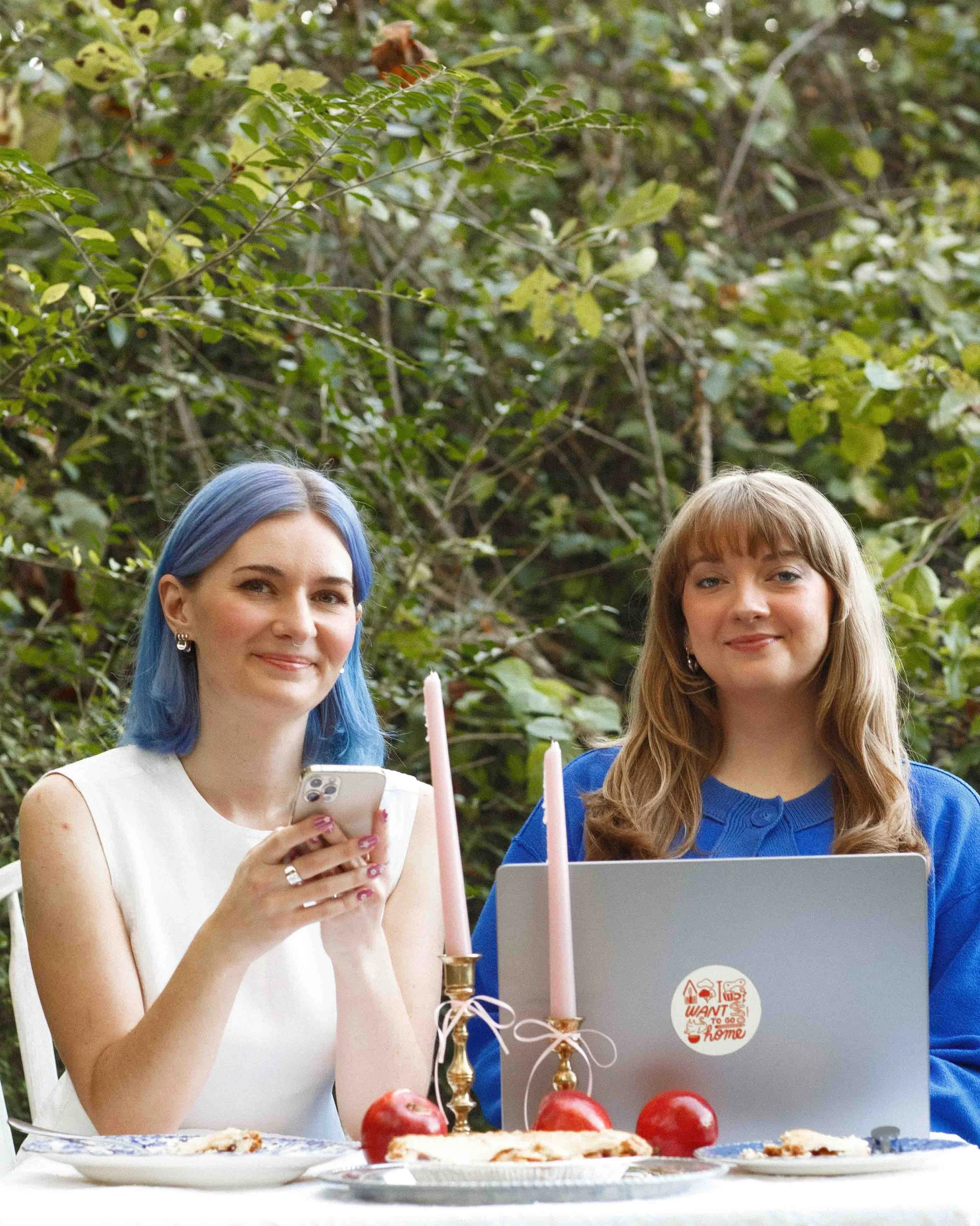 Foxtrot Cofounders Cass & Amanda posing and smiling sitting outdoors at a table with a laptop, pink candles, red apples and plates